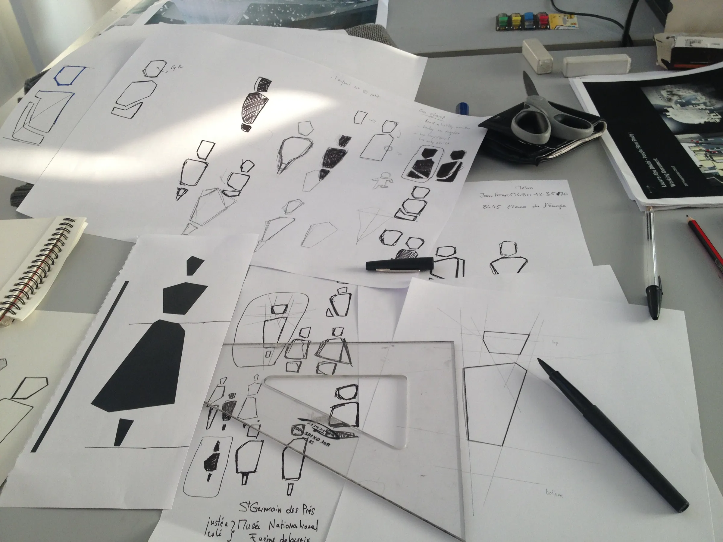 Desk covered with pen sketches and printouts of angular human pictogram studies, with a ruler and set square