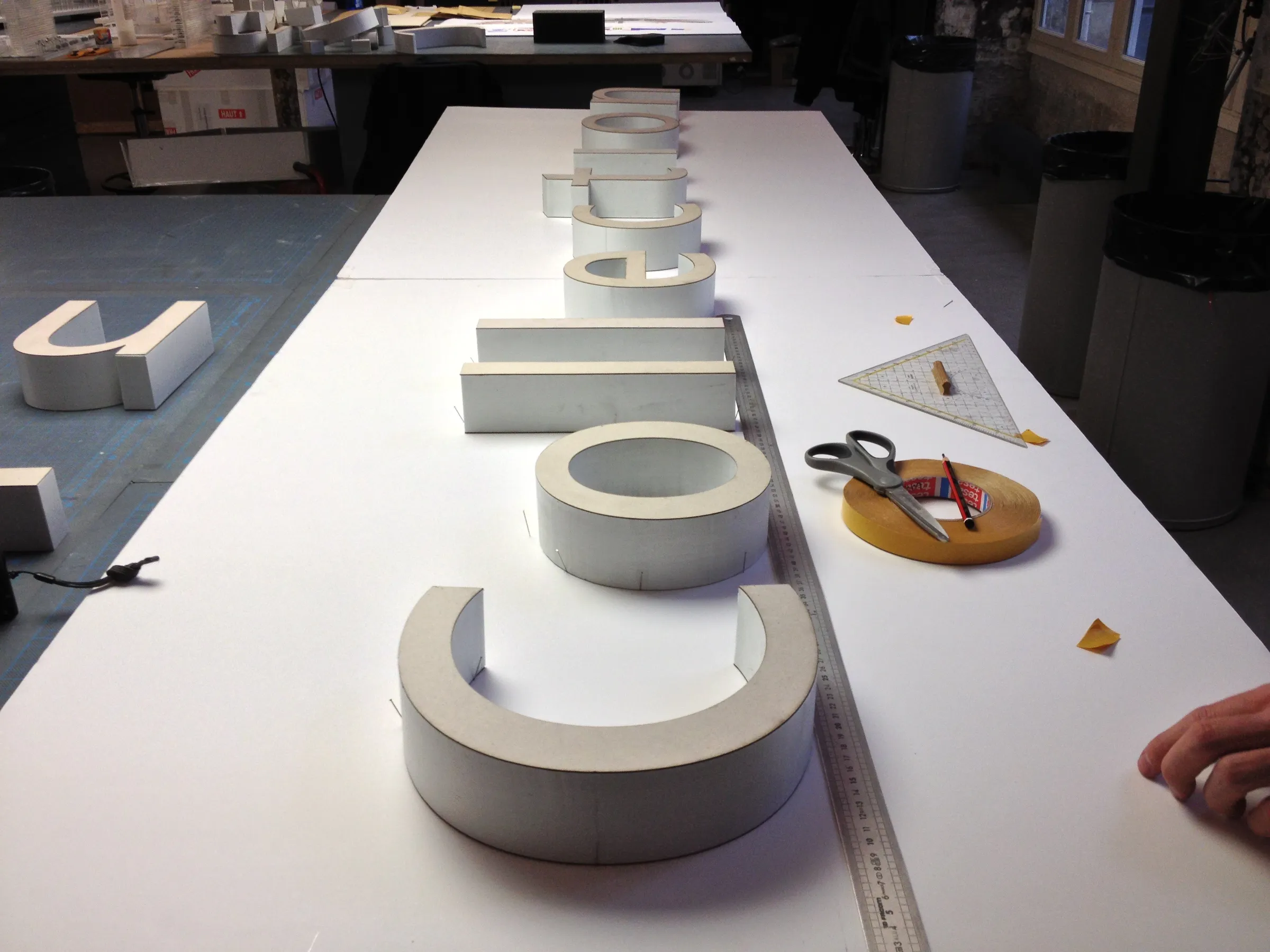 Row of white three-dimensional letters being assembled on a workshop table next to a ruler, tape and scissors